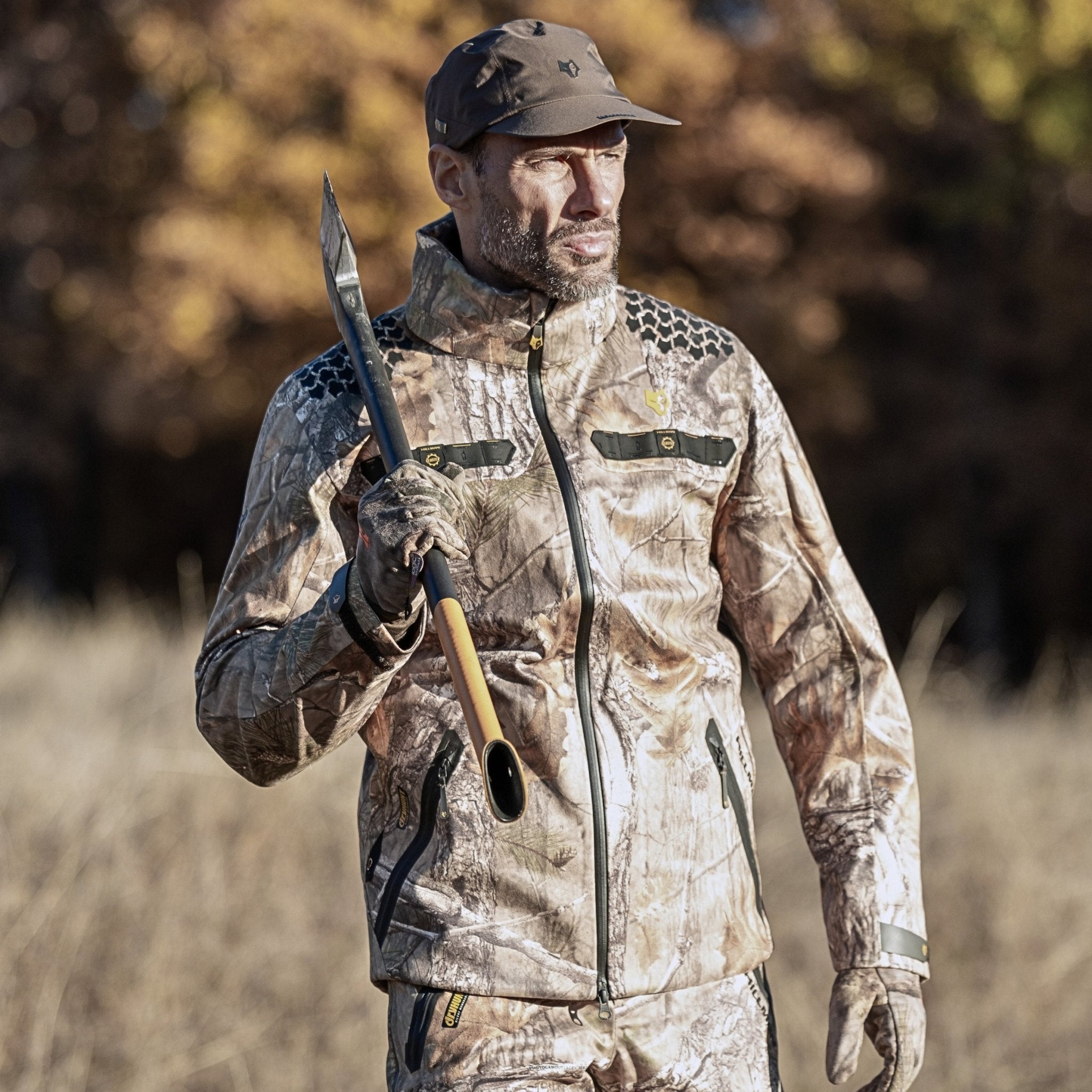 Ein Mann in Tarnkleidung und mit Mütze hält ein langes speerähnliches Werkzeug in der Hand. Er steht im Freien, im Hintergrund sind verschwommene Bäume zu sehen. Seine Hillman 5WL Camouflage Regenjacke hält ihn bei unerwarteten Regenschauern trocken.