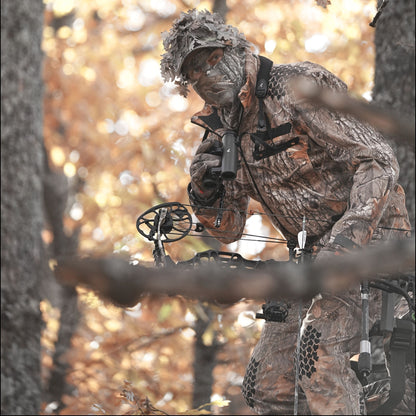 Eine Person in einer 6OL Winter Jagdjacke Camouflage von HILLMAN GEAR verschmilzt mit dem Herbstlaub, hält Pfeil und Bogen und blickt durch ein Fernglas in gedämpften Orange- und Brauntönen.