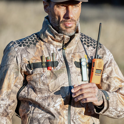 Eine Person in einer Hillman 5WL Camouflage Regenjacke – Wasserdichte Jagdjacke steht im Freien und hält ein tragbares Radio in der Hand. Die ultraleichte Jacke, auf der Vorderseite mit Schrotpatronen verziert, fügt sich nahtlos in die natürliche Umgebung ein. Mit einem konzentrierten Ausdruck sind sie für jedes Wetter bereit.