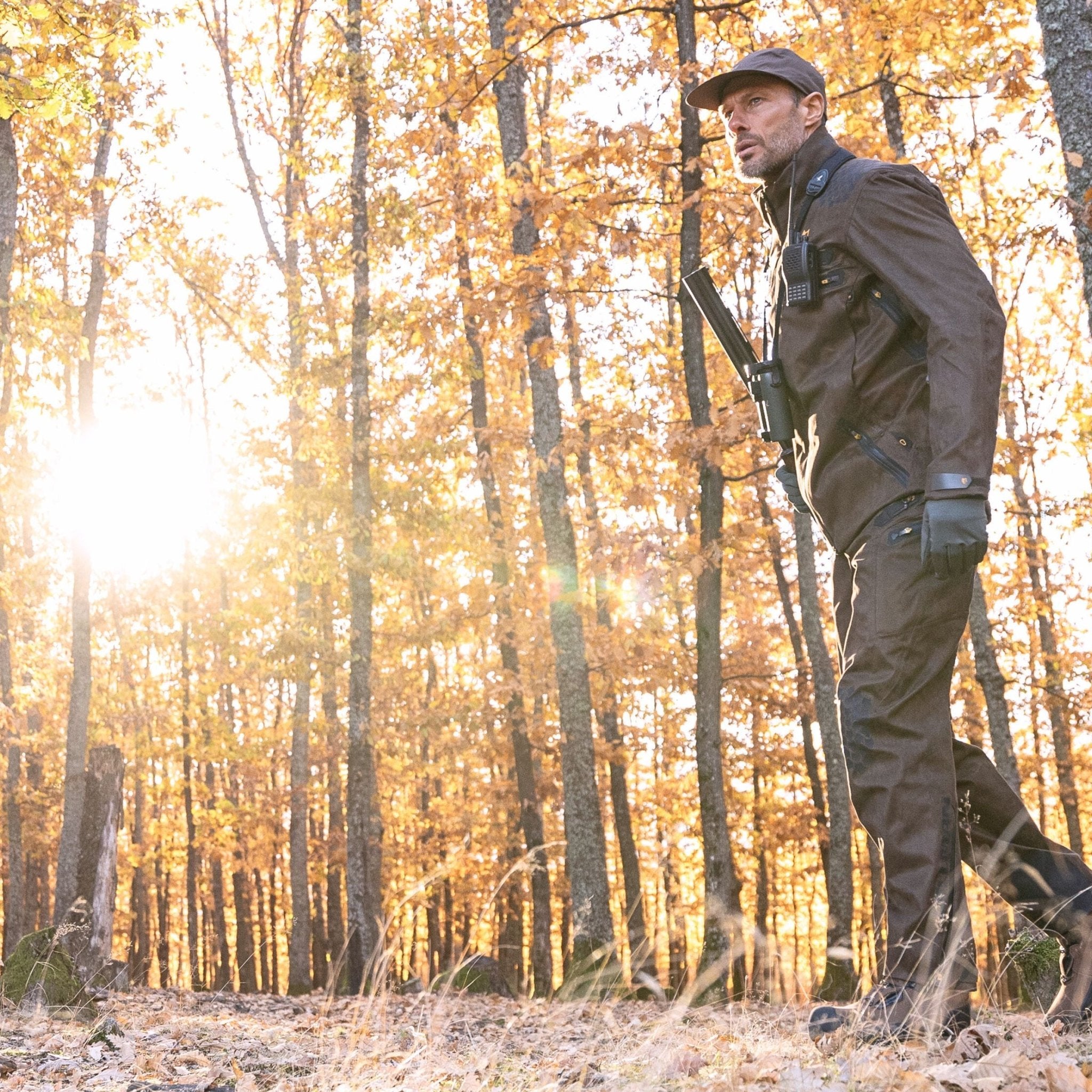 Sie schlendern durch einen sonnenbeschienenen Wald mit herbstlichem Laub und tragen ein braunes Outfit mit Mütze, Handschuhen und Hillmans 6OL Winter-Jagdhose. Mit Ferngläsern in der Hand sind sie offensichtlich in ihre Outdoor-Aktivitäten vertieft, während die Sonne durch die Bäume scheint.