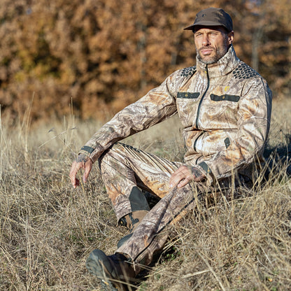 Ein Mann in einer Hillman 5WL Camouflage Regenjacke – einer ultraleichten, wasserdichten Jagdjacke – sitzt auf trockenem Gras. Seine passende Kleidung, bestehend aus Hose und Mütze, verschmilzt nahtlos mit den Herbstbäumen und ihren braunen Blättern im unscharfen Hintergrund.