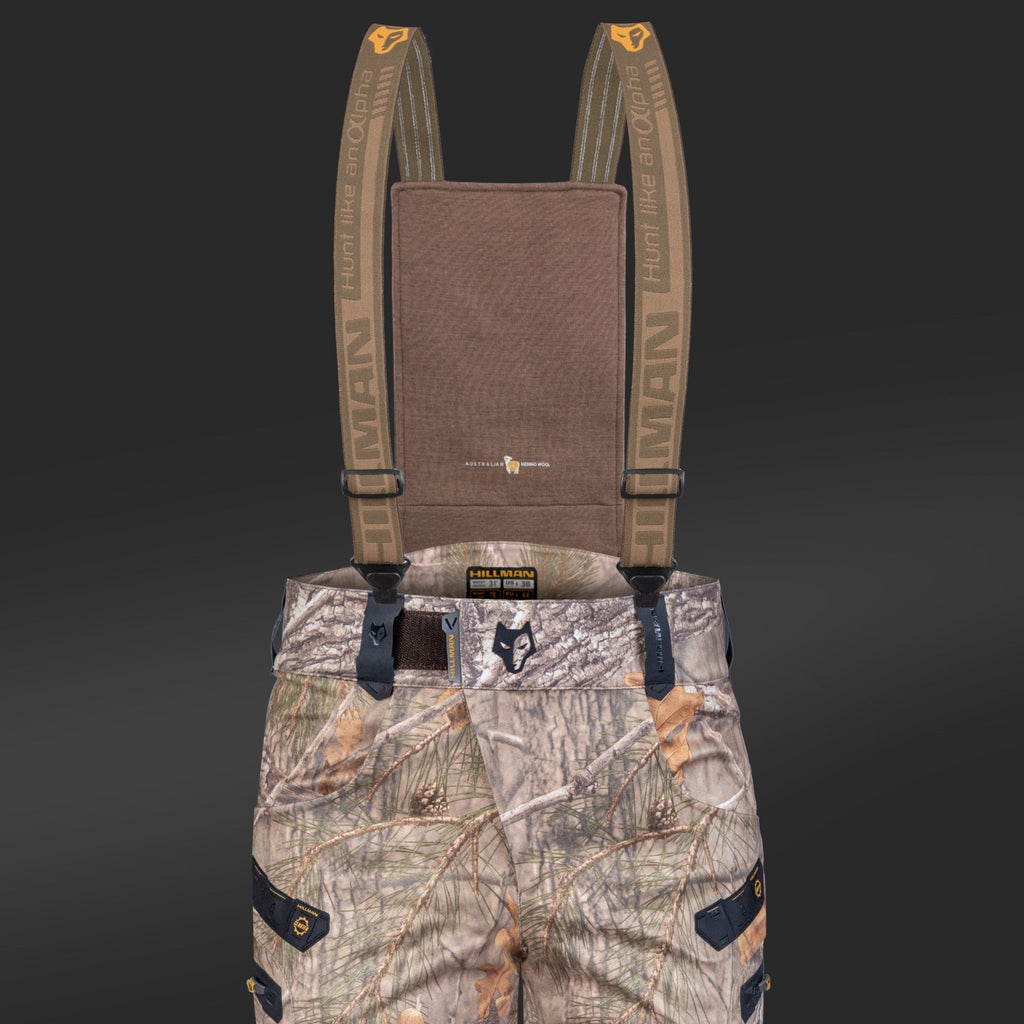 Die 6OL Winter-Jagdhose von Hillman mit Camouflage-Muster hat verstellbare Hosenträger, ist in Orange- und Brauntönen gehalten und trägt die Aufschrift „Hunters and Others“, „HUMAN“ und schwarze Taschen. Ihr robustes Design wird durch einen dunklen Hintergrund mit schwarzen Druckknöpfen ergänzt.