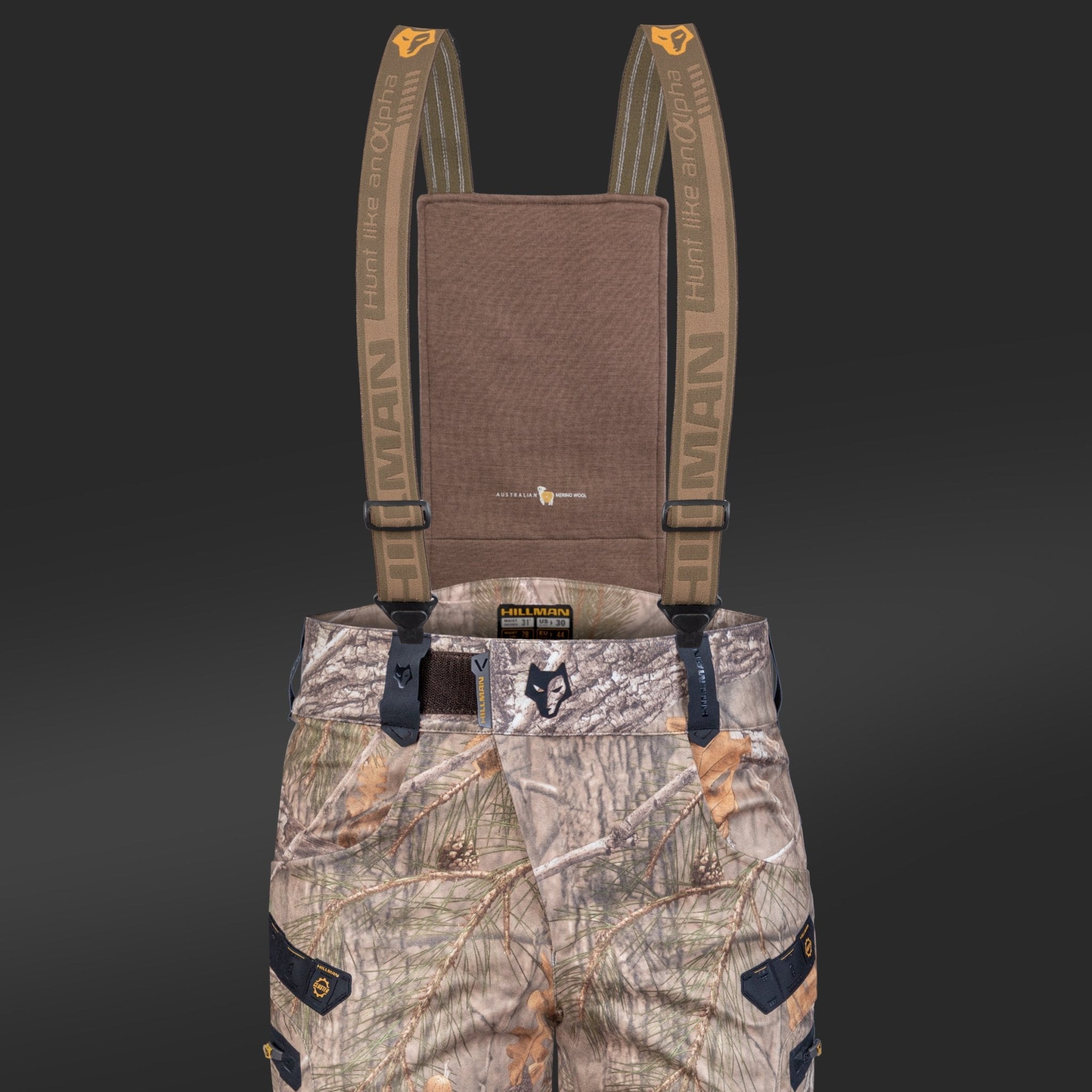 Die 6OL Winter-Jagdhose von Hillman mit Camouflage-Muster hat verstellbare Hosenträger, ist in Orange- und Brauntönen gehalten und trägt die Aufschrift „Hunters and Others“, „HUMAN“ und schwarze Taschen. Ihr robustes Design wird durch einen dunklen Hintergrund mit schwarzen Druckknöpfen ergänzt.