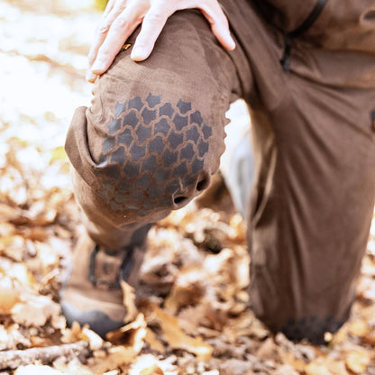 Eine Person kniet auf einer Waldlichtung und trägt Hillmans 6OL Winter-Jagdhose – mit Kniebesatz und braunen Stiefeln – und legt ihre Hand auf das Knie, bereit für die bevorstehende anspruchsvolle Wanderung durch die raue Wildnis.