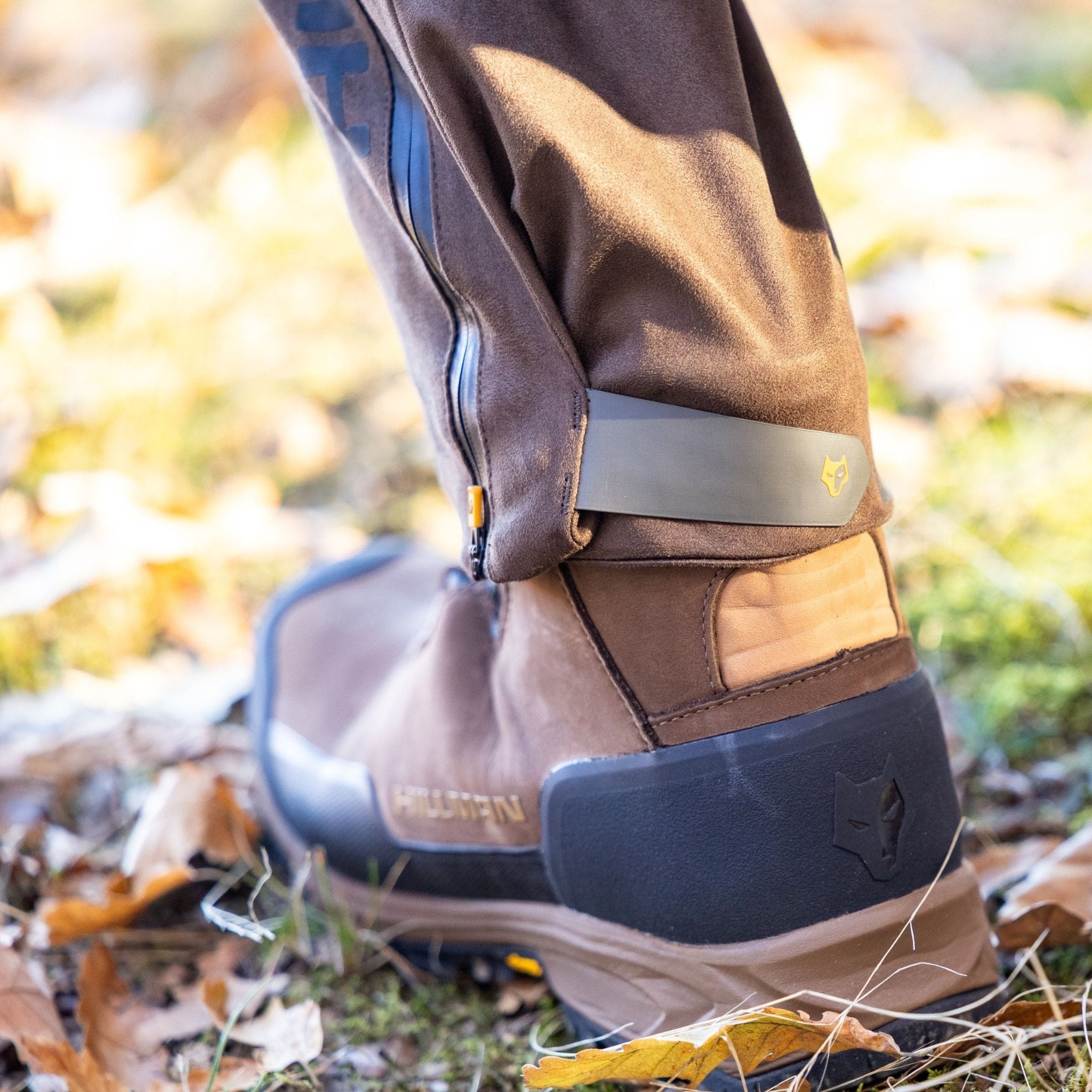 Nahaufnahme einer Person in braunen Wanderstiefeln und Hillmans 6OL Winter Jagdhose, einer wasserdichten, dornenfesten Trekkinghose für die Jagd im Freien. Die Hose ist über den Stiefeln teilweise mit einem Reißverschluss versehen, was ihr vielseitiges Design unterstreicht. Gefallene Blätter bedecken das Gras und lassen eine herbstliche Landschaft erahnen.