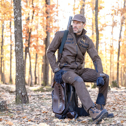 Ein Jäger, der Hillmans 6OL Winterjagdhose und eine braune Mütze trägt, sitzt auf einem Sack in einem Herbstwald. Mit gefallenen Blättern und hohen Bäumen mit orangefarbenem Laub fängt die Szene die lebendige Essenz eines Outdoor-Abenteuers ein.