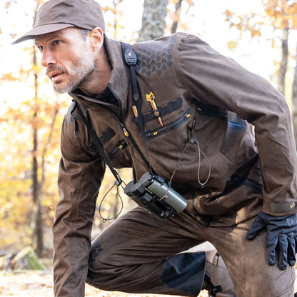 In einem Wald mit Herbstlaub hockt eine Person in einem braunen Outdoor-Outfit und trägt die 6OL Winter Jagdjacke - Wasserdichte Jacke von HILLMAN GEAR. Mit Mütze, Handschuhen und einem Fernglas um den Hals verkörpert sie Ruhe und Abenteuer.