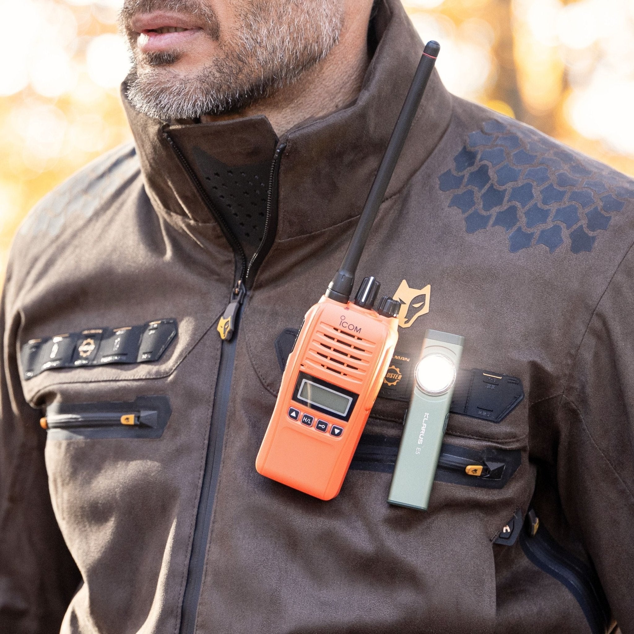 Eine Person trägt eine braune HILLMAN GEAR 6OL Winter-Jagdjacke mit mehreren Reißverschlüssen, dazu ein orangefarbenes Walkie-Talkie und eine daran befestigte Taschenlampe. Der sanft verschwommene Hintergrund lässt auf eine Außenkulisse mit warmer Beleuchtung schließen.