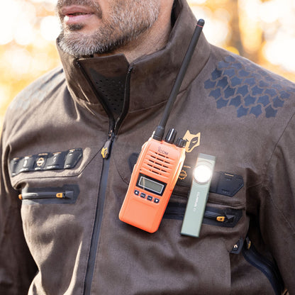 Eine Person trägt eine braune HILLMAN GEAR 6OL Winter-Jagdjacke mit mehreren Reißverschlüssen, dazu ein orangefarbenes Walkie-Talkie und eine daran befestigte Taschenlampe. Der sanft verschwommene Hintergrund lässt auf eine Außenkulisse mit warmer Beleuchtung schließen.