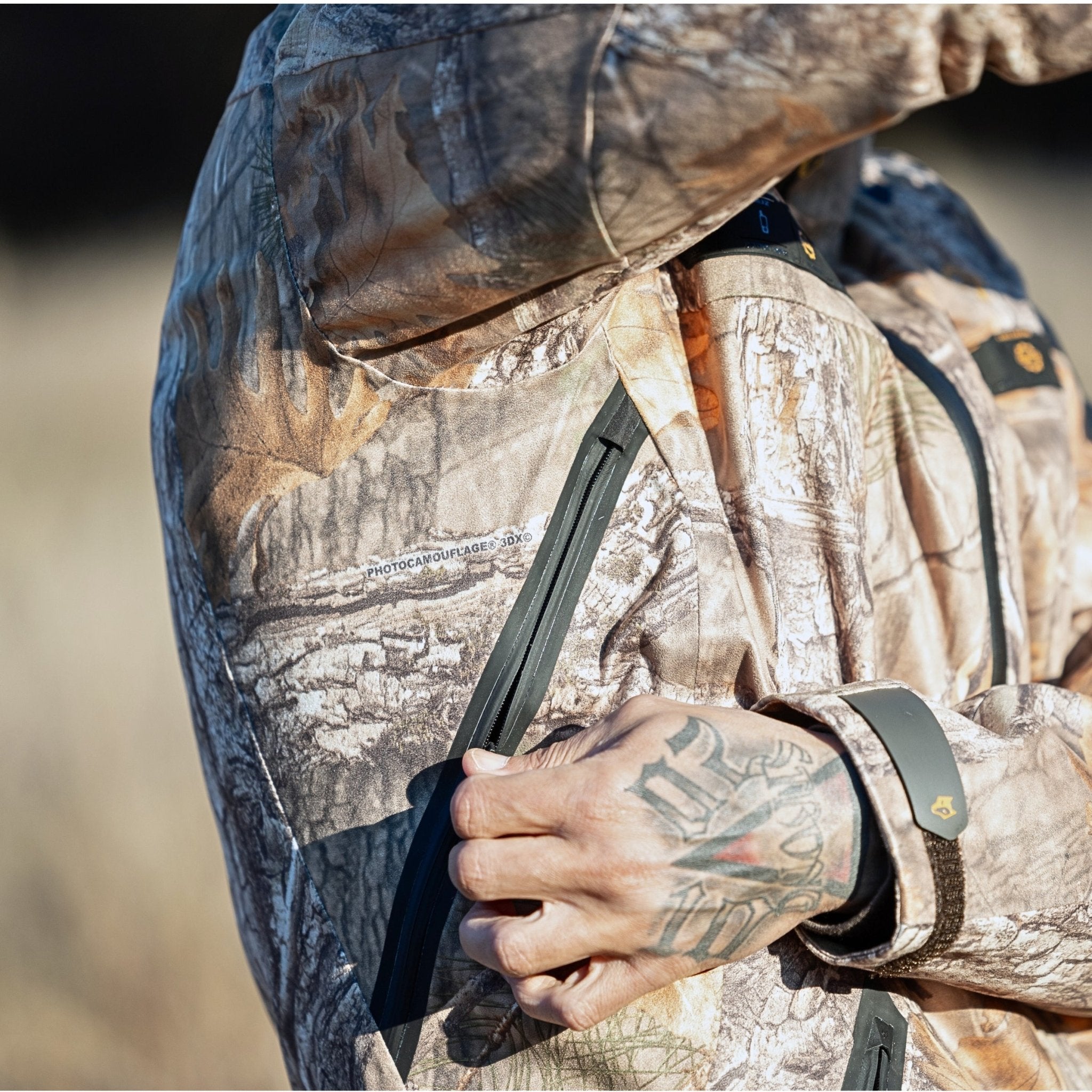 Eine tätowierte Hand passt den Ärmelreißverschluss einer Hillman 5WL Camouflage Regenjacke an – einer ultraleichten, wasserdichten Jagdjacke. Die Jacke verschmilzt nahtlos mit dem trockenen Gras und dem verschwommenen dunklen Hintergrund.