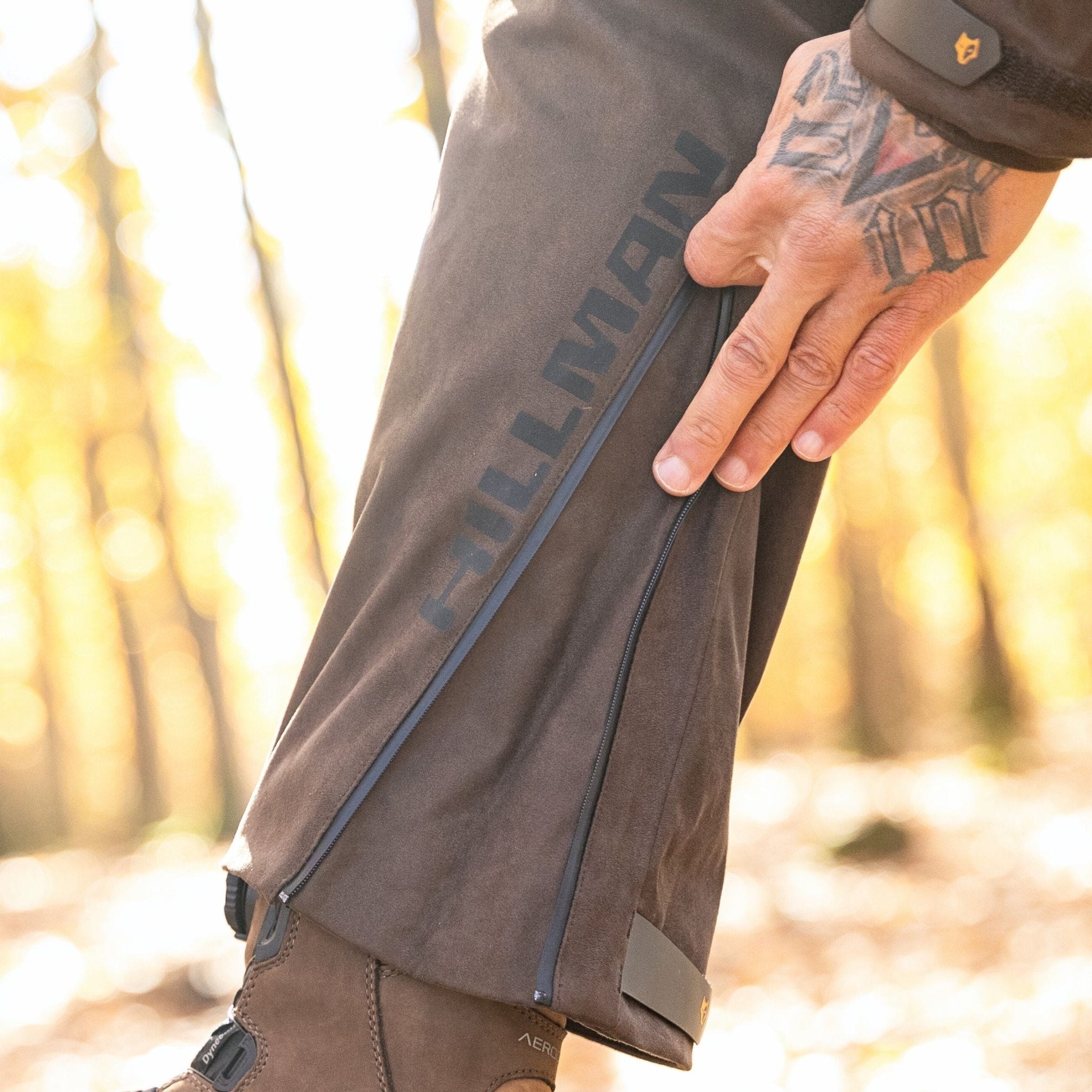 Eine Person mit einer tätowierten Hand passt den seitlichen Reißverschluss ihrer braunen 6OL Winter-Jagdhose von Hillman an. Sie steht an einem sonnigen Tag im Freien, mit verschwommenen Bäumen im Hintergrund, und trägt robuste braune Stiefel, die ihren robusten Stil unterstreichen.