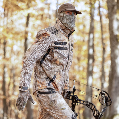 Eine Person in einer 6OL Winter-Jagdjacke von HILLMAN GEAR schreitet durch einen Herbstwald, mit passender Gesichtsbedeckung und Handschuhen. Sie hält einen Compound-Bogen in der Hand und ist bereit für die Jagd.