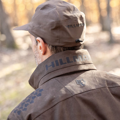 Eine Person in einer braunen, atmungsaktiven 6OL Winter-Jagdjacke und Mütze steht draußen in einem sonnenbeschienenen Waldstück. Beide sind mit der Marke „HILLMAN GEAR“ und einem Wolfslogo vor verschwommenen Bäumen versehen und zeigen die innovativen Materialien, die in dieser wasserdichten Jagdjacke verwendet werden.