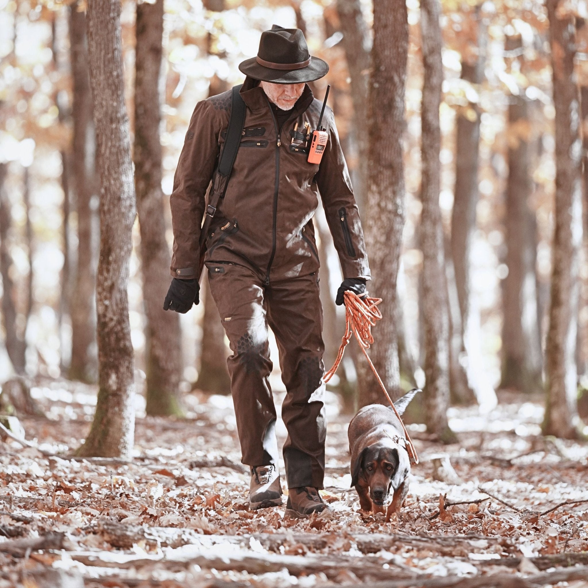 Eine Person in braunem Outfit und Hut geht mit einem Hund an der Leine durch einen grünen Wald. Sie trägt die strapazierfähige Hillman 6OL Winter Jagdhose – wasserdichte dornenfeste Trekkinghose für die Jagd. Sie trägt ein Funkgerät und scheint sich auf ihren Spaziergang zwischen hohen Bäumen zu konzentrieren.