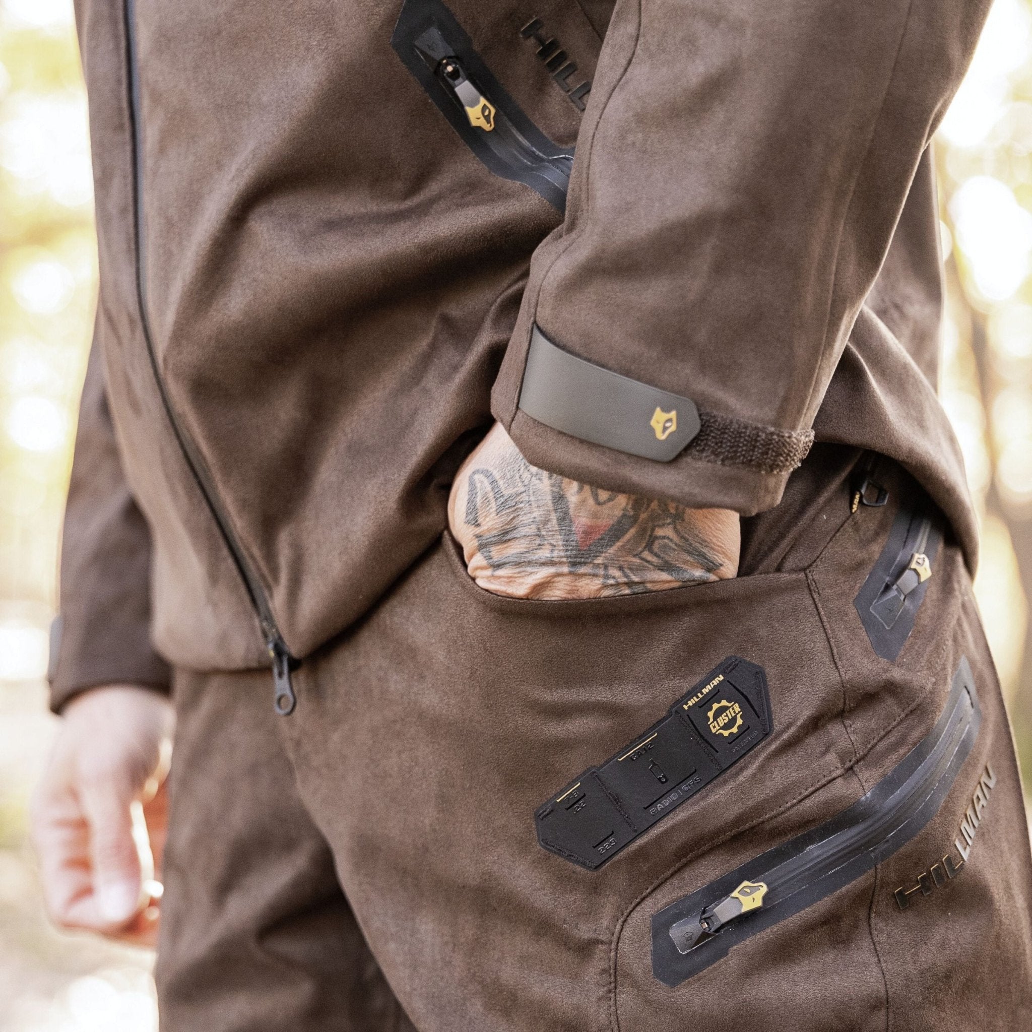 Eine Person in einem braunen Wildlederoutfit mit aufwendigen Flickendetails und Tattoos steht mit der Hand in der Tasche draußen und hebt das Design der Jacke hervor, das an eine robuste Hillman 6OL Winter-Jagdhose erinnert.