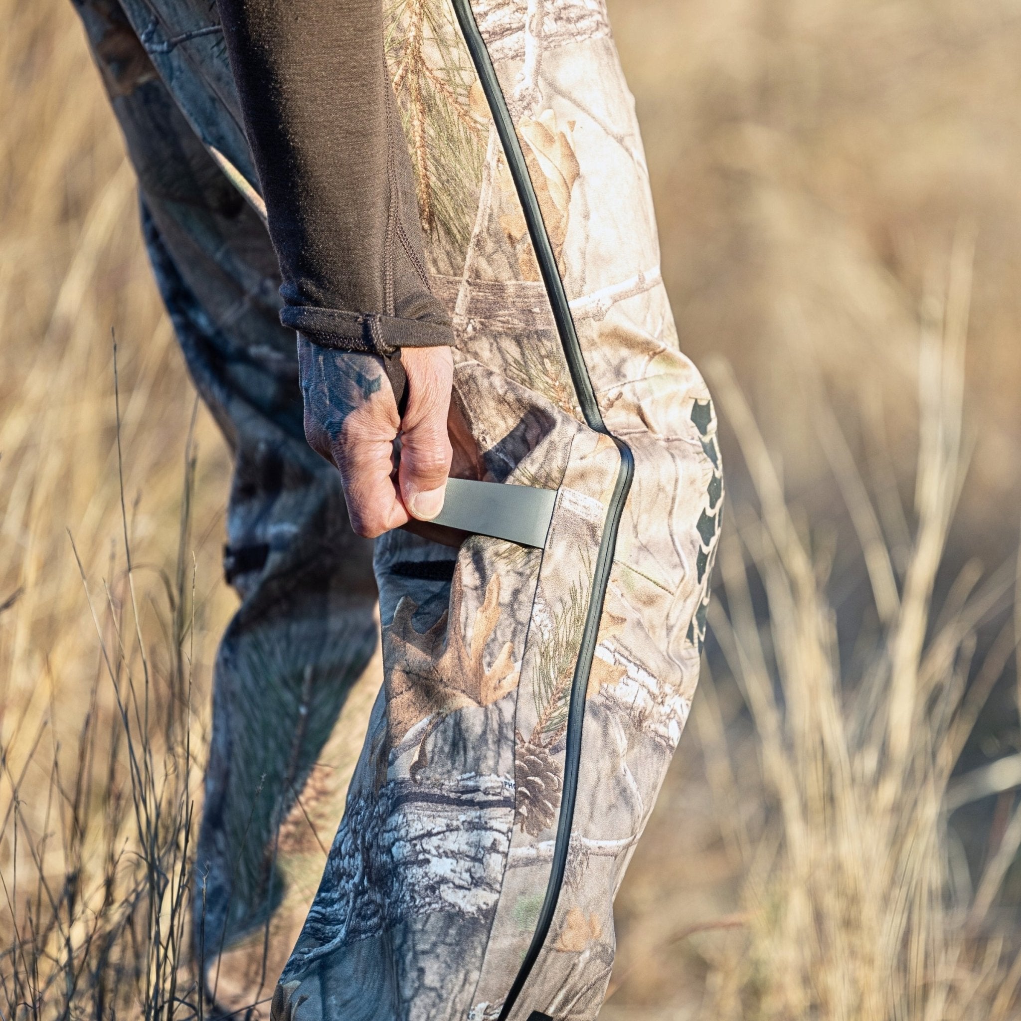 Eine Hand greift nach dem Reißverschluss einer Hillman 5WL Camouflage Regenhose, einer ultraleichten, wasserdichten Jagdhose. In ein langärmeliges Hemd gekleidet steht die Person zwischen trockenem Gras und präsentiert die komplizierten Details dieser funktionalen Ausrüstung.